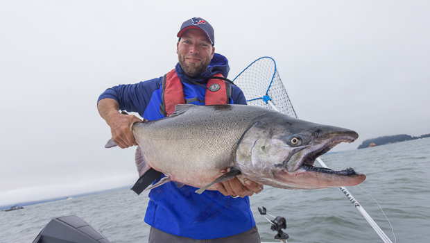 Smiling man in a blue jacket and ball cap holding a Chinook salmon with its mouth open. Fishing at the Buoy 10 fishery out of Astoria, Oregon.